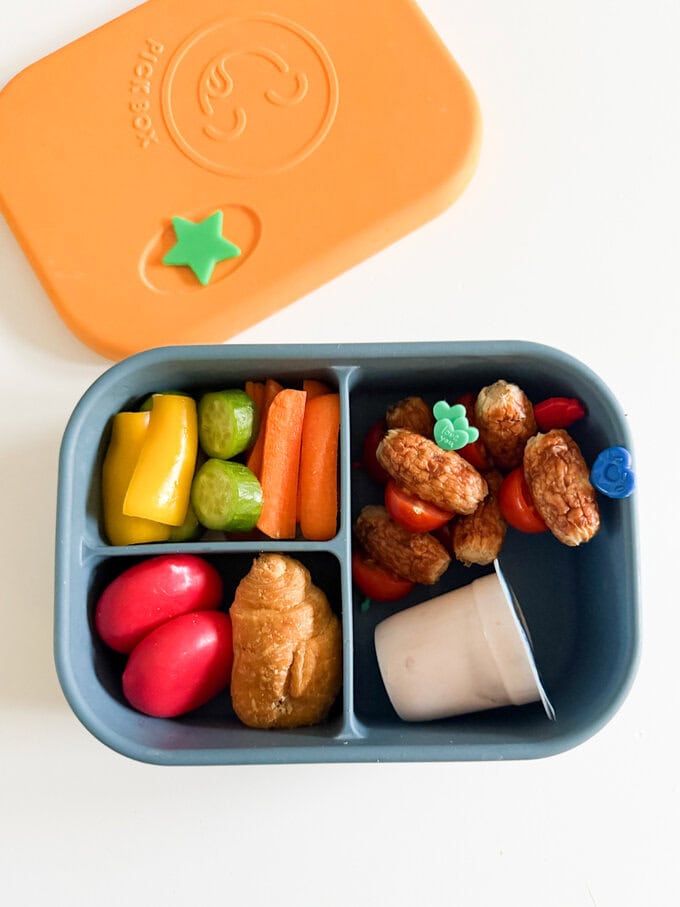 Sausage & Tomato skewers in a blue and yellow bento style lunchbox with chopped veggies, mini cheese portions, a mini croissant and a yogurt.