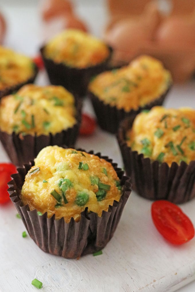 Pea & Sweetcorn Egg Muffins in brown paper muffin cases on a white chopping board with chopped cherry tomatoes and chives.