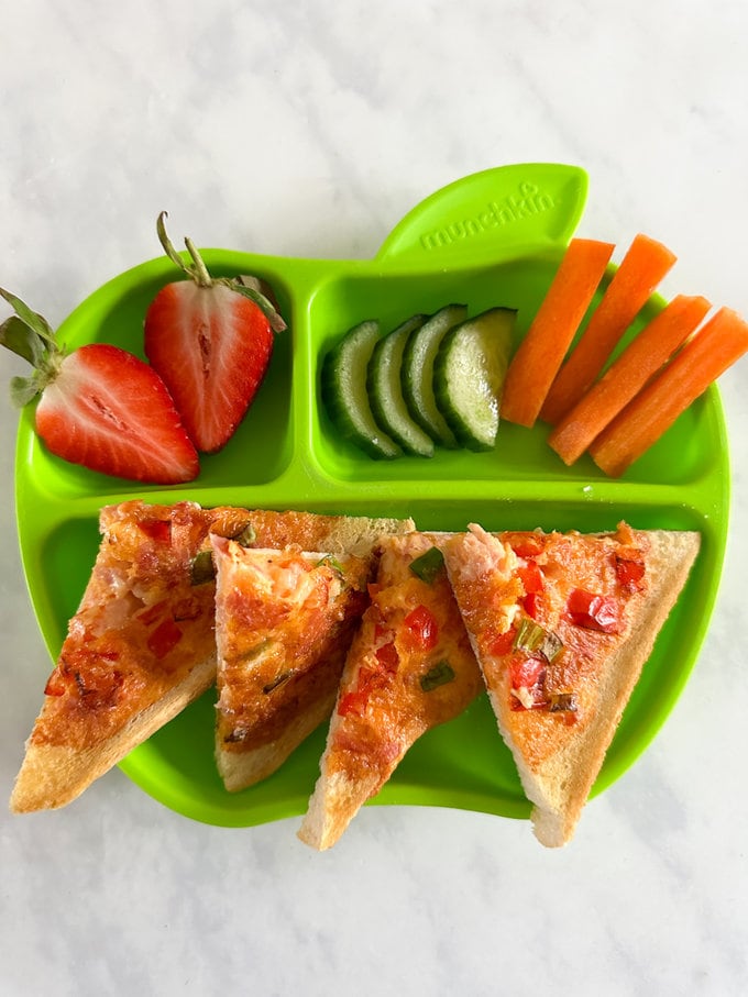 Airfryer omelette toast presented on a green apple shaped plastic child's plate and served with fresh chopped strawberries, carrot and cucumber.