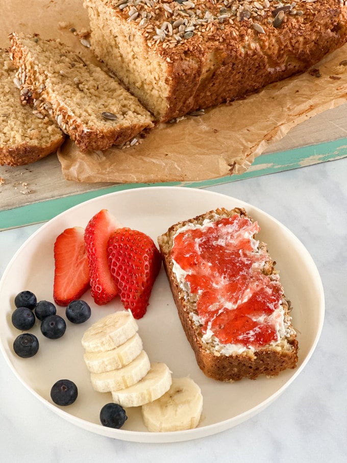 A thick slice of Porridge Bread spread with butter and jam served on a white side plate. With sliced banana, blueberries and slices strawberries.
